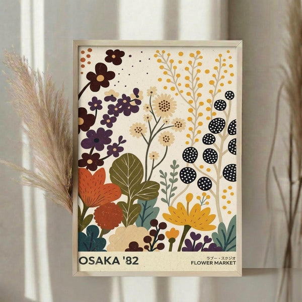 May include: Framed botanical poster art featuring stylized flowers in warm earth tones including rust orange, deep purple, golden yellow, and olive green against cream background. Modern minimalist illustration style with geometric florals, berries, and leaves. Natural wood frame displayed on white surface with dried wheat stems beside it, soft natural lighting from window in background.
