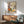 May include: Framed botanical poster with colorful stylized flowers and plants in earth tones - orange, purple, yellow, and green blooms against cream background. Displayed above mid-century modern walnut credenza with sliding doors and ceramic table lamp. Sage green upholstered armchair with wooden frame sits nearby on light hardwood floors. Soft natural lighting creates shadows on white wall.
