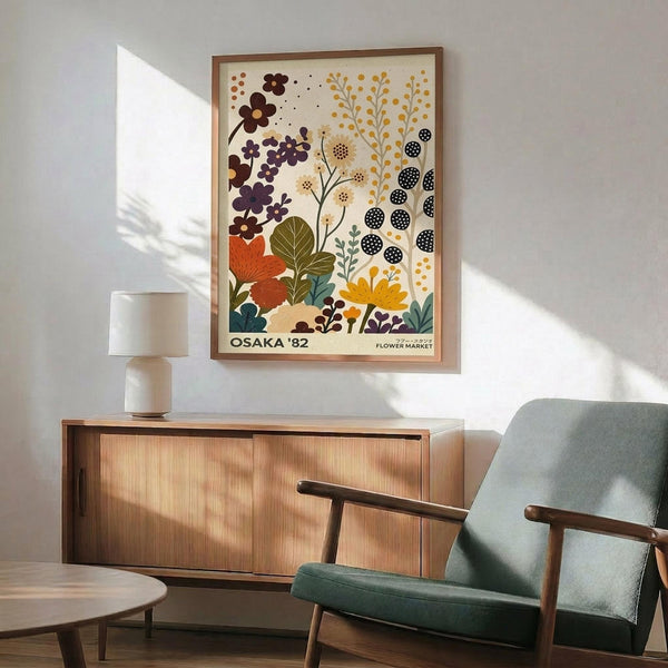 May include: Framed botanical poster with colorful stylized flowers and plants in earth tones - orange, purple, yellow, and green blooms against cream background. Displayed above mid-century modern walnut credenza with sliding doors and ceramic table lamp. Sage green upholstered armchair with wooden frame sits nearby on light hardwood floors. Soft natural lighting creates shadows on white wall.