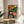 May include: Framed botanical print with stylized flowers and leaves in vibrant orange, coral, yellow, and green colors against dark background, displayed on round wooden side table next to beige upholstered chair. Natural sunlight streams across white walls creating soft shadows. Small plant visible in corner adds organic touch to modern interior setting.