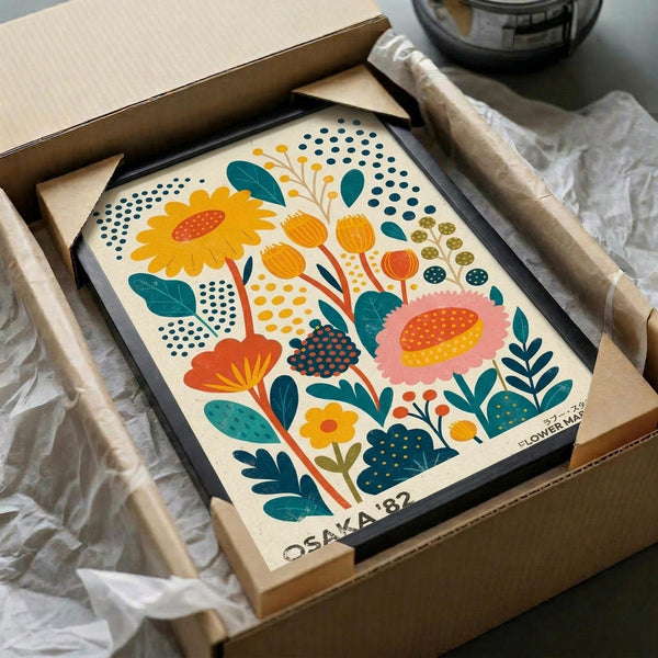 Colorful floral art print in black frame being unboxed from cardboard packaging with tissue paper. Features stylized flowers in orange, pink, yellow and teal with geometric patterns and dotted details in folk art style. Print shows sunflowers, leaves and botanical elements on cream background. Frame sits in brown shipping box surrounded by white protective wrapping.