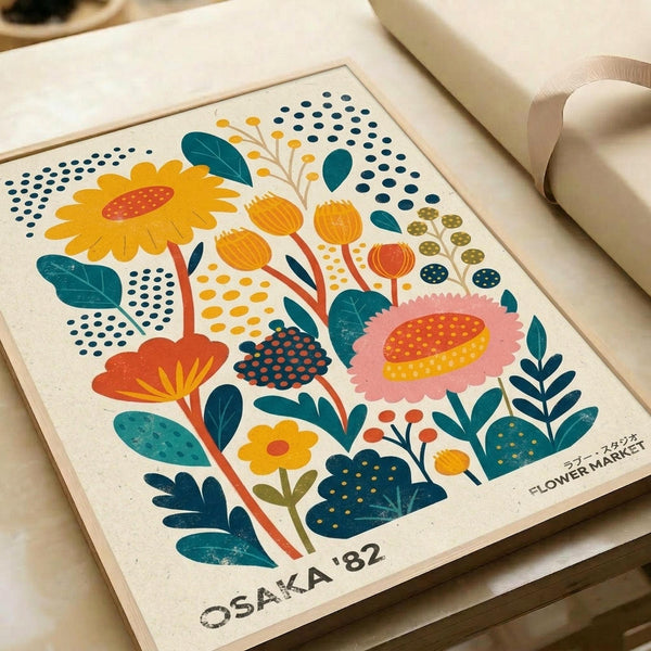 Colorful retro-style botanical illustration featuring stylized flowers in orange, pink, and yellow with teal leaves and dotted patterns. The artwork shows daisies, abstract blooms, and foliage arranged in a folk art style on cream paper. Text reads "OSAKA '82" and "FLOWER MARKET" in bottom corners. The print sits on a wooden surface next to rolled paper tubes.