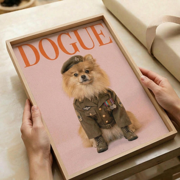 May include: Hands holding a framed poster print featuring a fluffy Pomeranian dog dressed as a military general in olive green uniform with medals, beret, and boots against a pink background. The word "DOGUE" appears in large orange letters at the top. The artwork is displayed in a light wood frame on white bedding with cream ribbon visible nearby.