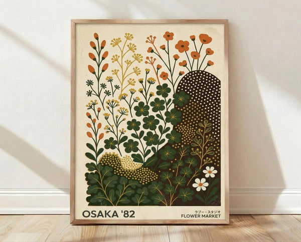 May include: Framed botanical poster print leaning against white wall on light wooden floor. Cream-colored artwork features stylized wildflowers and plants in earthy tones of orange, green, and gold with abstract dotted shapes. Text reads "OSAKA '82" and "FLOWER MARKET" in both English and Japanese characters. Natural wood frame, soft lighting creates gentle shadows on textured white wall.