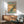 May include: Framed poster featuring a blue and green peacock perched on cherry blossom branches against warm golden sky, displayed above wooden sideboard with white ceramic lamp and gray upholstered armchair in modern living room with natural lighting