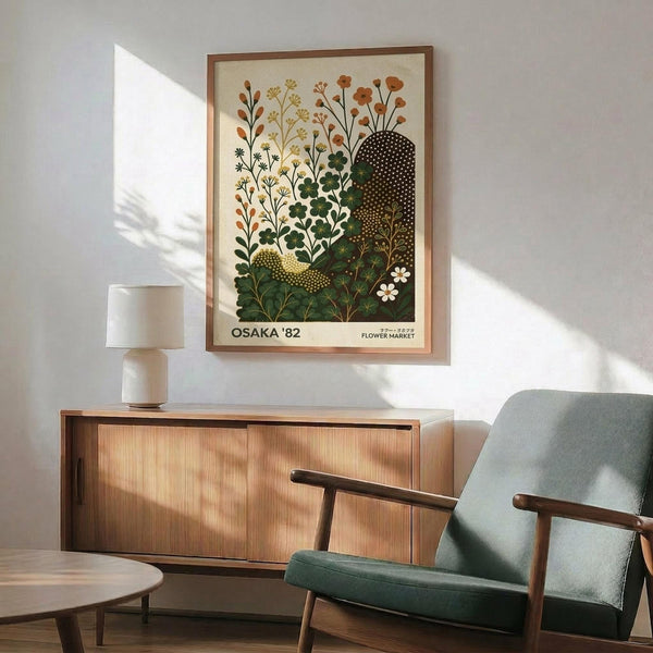May include: Botanical poster print featuring stylized wildflowers and plants in earth tones of orange, green, and brown, displayed in thin wooden frame above mid-century modern credenza. Sage green upholstered chair with wood arms sits beside round coffee table, white ceramic table lamp, against white wall with natural sunlight shadows.