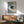 May include: Framed poster print featuring a black cat with yellow eyes curled up on blue and white striped bedding, holding a coffee cup. The artwork has a minimalist, illustrated style in a thin wooden frame. Room setting shows mid-century modern furniture including a sage green upholstered armchair with wooden arms, walnut credenza, white ceramic table lamp, and round coffee table. Natural sunlight creates geometric shadows on the white wall.