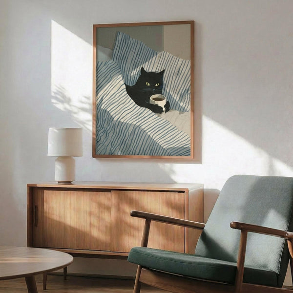 May include: Framed poster print featuring a black cat with yellow eyes curled up on blue and white striped bedding, holding a coffee cup. The artwork has a minimalist, illustrated style in a thin wooden frame. Room setting shows mid-century modern furniture including a sage green upholstered armchair with wooden arms, walnut credenza, white ceramic table lamp, and round coffee table. Natural sunlight creates geometric shadows on the white wall.