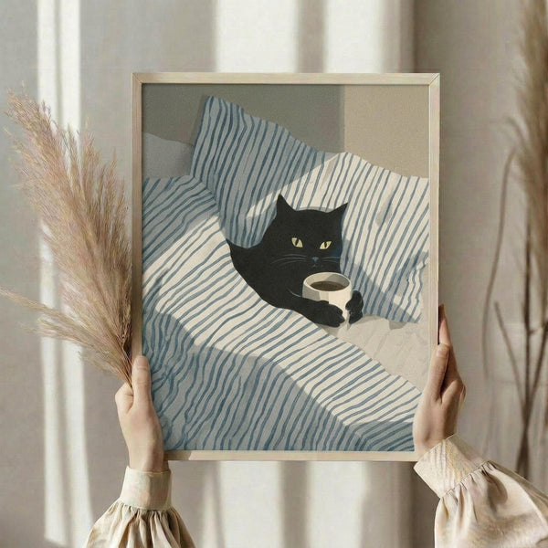 May include: Person holding framed artwork showing black cat with yellow eyes holding white coffee cup, resting on blue and white striped bedding. One hand also holds dried pampas grass stems. Light wooden frame against soft neutral bedroom setting with natural window light.
