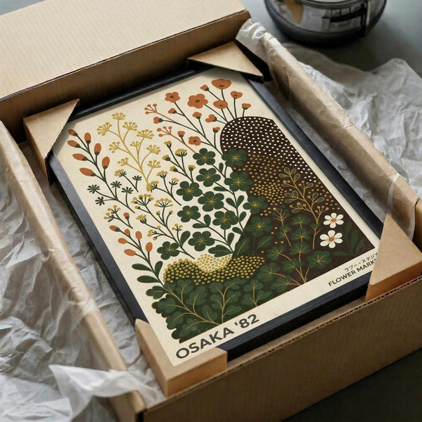 May include: Framed botanical art print in black frame, nestled in cardboard box with white tissue paper. The artwork features stylized flowers and plants in earthy tones - rust orange blooms, golden yellow stems, deep green foliage, and white daisies arranged in decorative patterns against cream background. Print shows "OSAKA '82" and "FLOWER MARKET" text at bottom. Wooden dowels and dark glass jar visible in background on white fabric surface.