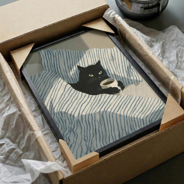 May include: Framed print showing black cat curled up on blue and white striped bedding holding a white coffee cup, displayed in black frame being unpacked from cardboard shipping box with protective wrapping on white surface.