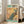May include: Framed vintage-style poster featuring a vibrant peacock perched on cherry blossom branches against a golden yellow background. The peacock displays detailed blue and teal plumage with ornate tail feathers. Text reads "1967 JAPAN WEEK" in green and black lettering. The wooden-framed artwork leans against a white wall next to a beige upholstered chair in a bright, minimalist living room with large windows and natural lighting.