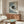 May include: Framed poster of black cat with yellow eyes holding coffee cup on blue and white striped bedding, displayed above wooden credenza in modern living room with white curved chair, glass coffee table, and table lamp