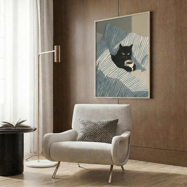 May include: Framed poster print featuring a black cat with yellow eyes lying on blue and white striped bedding, holding a coffee cup. Modern minimalist artwork displayed in a contemporary living room setting with beige armchair, geometric throw pillow, brass floor lamp, and wood paneled wall. Soft natural lighting from nearby window creates warm ambiance.