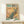 May include: Framed vintage-style poster featuring a detailed peacock perched on cherry blossom branches against a golden sunset sky with mountain backdrop. The peacock displays vibrant teal and blue plumage with distinctive eye-spot tail feathers. Text reads "1967 Japan Week" in large green and black letters at top, with Japanese characters and "Heritage Paintings" details below. Wooden frame sits on light wood furniture next to a small white wrapped gift with evergreen sprig.
