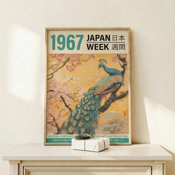 May include: Framed vintage-style poster featuring a detailed peacock perched on cherry blossom branches against a golden sunset sky with mountain backdrop. The peacock displays vibrant teal and blue plumage with distinctive eye-spot tail feathers. Text reads "1967 Japan Week" in large green and black letters at top, with Japanese characters and "Heritage Paintings" details below. Wooden frame sits on light wood furniture next to a small white wrapped gift with evergreen sprig.