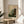 May include: A framed botanical art print displayed on a round wooden side table next to a beige upholstered chair. The artwork features stylized wildflowers and plants in warm oranges, yellows, and greens against a cream background, with dotted black silhouettes creating contrast. Natural sunlight streams across the white walls, casting soft shadows from a potted plant visible in the corner. The print has a thin wooden frame and includes text at the bottom.