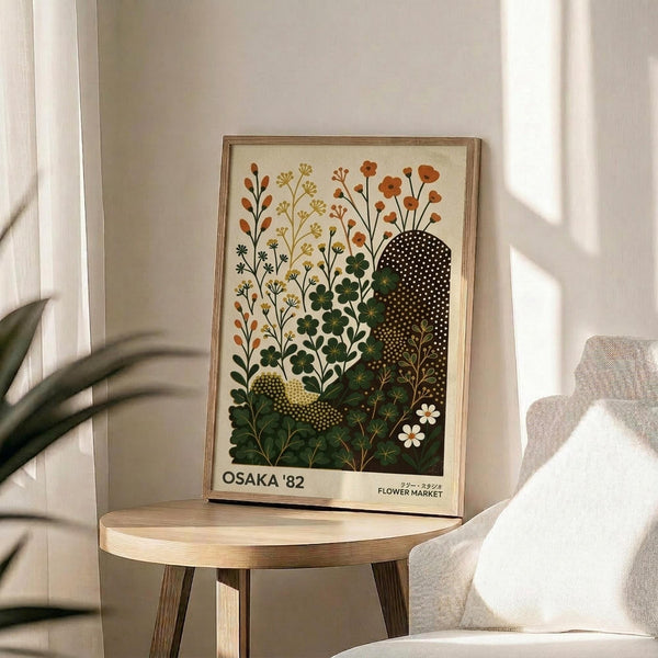 May include: A framed botanical art print displayed on a round wooden side table next to a beige upholstered chair. The artwork features stylized wildflowers and plants in warm oranges, yellows, and greens against a cream background, with dotted black silhouettes creating contrast. Natural sunlight streams across the white walls, casting soft shadows from a potted plant visible in the corner. The print has a thin wooden frame and includes text at the bottom.
