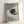 May include: Framed poster print leaning against white wall showing black cat with yellow-green eyes sitting on blue and white striped bedding, holding small coffee cup. Light wood frame, soft natural lighting, cozy bedroom scene with rumpled striped sheets and pillows.