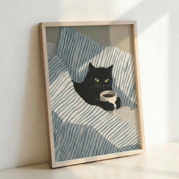 May include: Framed poster print leaning against white wall showing black cat with yellow-green eyes sitting on blue and white striped bedding, holding small coffee cup. Light wood frame, soft natural lighting, cozy bedroom scene with rumpled striped sheets and pillows.