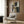 May include: Abstract artwork with geometric shapes in navy blue, gold, beige and white tones, displayed in light wooden frame on wood-paneled wall. Modern cream armchair with geometric patterned pillow sits below artwork. Brass floor lamp and dark round side table with open book complete the contemporary room setting with natural lighting from window.