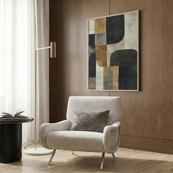May include: Abstract artwork with geometric shapes in navy blue, gold, beige and white tones, displayed in light wooden frame on wood-paneled wall. Modern cream armchair with geometric patterned pillow sits below artwork. Brass floor lamp and dark round side table with open book complete the contemporary room setting with natural lighting from window.