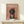 May include: Framed poster print displaying a dachshund wearing a pink chef's hat and heart-patterned apron on a dusty rose background with "DOGUE" text in cream letters at top. The golden-framed artwork sits on a light wooden shelf next to a small white wrapped gift with greenery sprig, against a white wall with soft natural lighting.