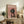 May include: Framed poster featuring a black and tan dachshund wearing a pink chef's hat and floral apron on a dusty rose background with white "DOGUE" text at top, displayed on rustic wooden console table alongside fern plant, books, ceramic vase, and brown leather chair in cozy living room setting