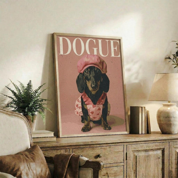May include: Framed poster featuring a black and tan dachshund wearing a pink chef's hat and floral apron on a dusty rose background with white "DOGUE" text at top, displayed on rustic wooden console table alongside fern plant, books, ceramic vase, and brown leather chair in cozy living room setting