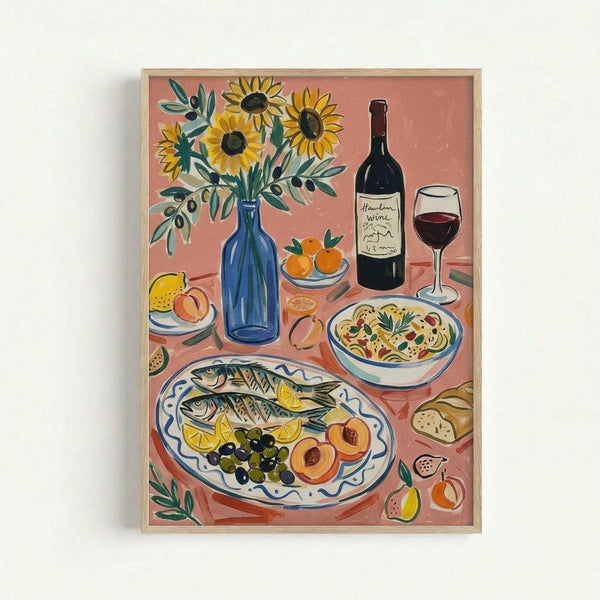 A framed still life illustration featuring a Mediterranean feast on a coral pink background. The artwork shows sunflowers in a blue bottle vase, grilled fish with lemon slices and olives on a decorative white platter, a bowl of pasta, wine bottle and glass, fresh oranges, peaches, and bread scattered across the composition. The painting style is folk art-inspired with bold colors and simple forms in a light wooden frame.