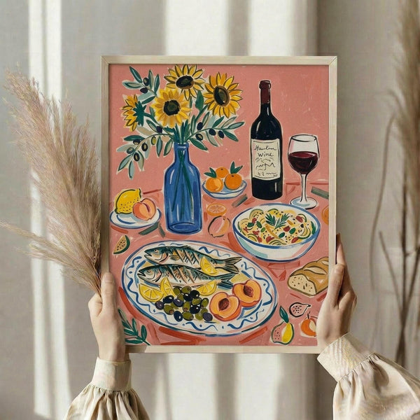 May include: Hands holding a framed still life illustration featuring sunflowers in a blue vase, grilled fish with lemon and olives on a decorative plate, wine bottle and glass, pasta bowl, and fresh fruit on a coral pink background. The artwork has a Mediterranean aesthetic with warm colors and is displayed against white curtains with dried pampas grass visible to the left.