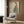 May include: Framed Japanese-style artwork showing woman in green striped kimono with coral floral sleeves holding persimmon, displayed on wood-paneled wall above beige upholstered chair with geometric patterned pillow, brass floor lamp, and dark round side table with open book in modern living room setting