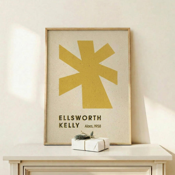 May include: Framed art print featuring a golden yellow abstract star or asterisk shape on cream background, displayed on light wood console table against white wall. Small wrapped gift box with evergreen sprig sits beside the natural wood frame. Minimalist Scandinavian-style interior setting with clean lines and neutral tones.