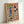 May include: Framed art print leaning against white wall showing colorful still life illustration with coral pink background. Features blue vase with yellow sunflowers, wine bottle and glass, decorative plate with whole fish, lemons and olives, bowls of pasta and fruit, olive branches. Painted in vibrant Mediterranean style with natural wood frame.
