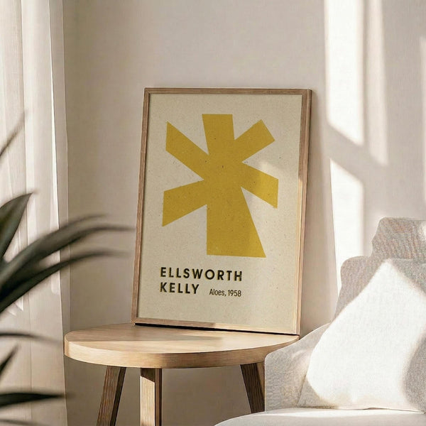 May include: Framed poster featuring a large yellow star-like geometric shape on cream background with black text reading "ELLSWORTH KELLY Aloes, 1958" below. Natural wood frame sits on round wooden side table next to beige upholstered chair with white pillow. Green plant leaves visible on left, soft natural lighting creates window shadows on white wall behind.