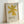 Framed minimalist art print featuring a bold yellow star-like shape with eight pointed rays against a cream textured background. The geometric form is positioned in the upper portion with "ELLSWORTH KELLY" and "Aloes, 1958" printed in dark text below. Displayed in a light wood frame leaning against a white wall, creating soft shadows.