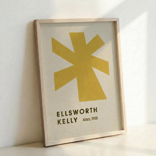 Framed minimalist art print featuring a bold yellow star-like shape with eight pointed rays against a cream textured background. The geometric form is positioned in the upper portion with "ELLSWORTH KELLY" and "Aloes, 1958" printed in dark text below. Displayed in a light wood frame leaning against a white wall, creating soft shadows.
