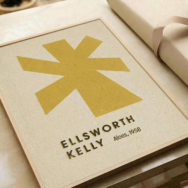A minimalist art print featuring a large golden yellow asterisk or star-like symbol centered on cream-colored textured paper. Below the symbol, black text reads "ELLSWORTH KELLY" and "Aloes, 1958". The print is displayed in a simple light wood frame alongside rolled paper tubes on a neutral surface, showing the artwork's clean geometric design and warm color palette.