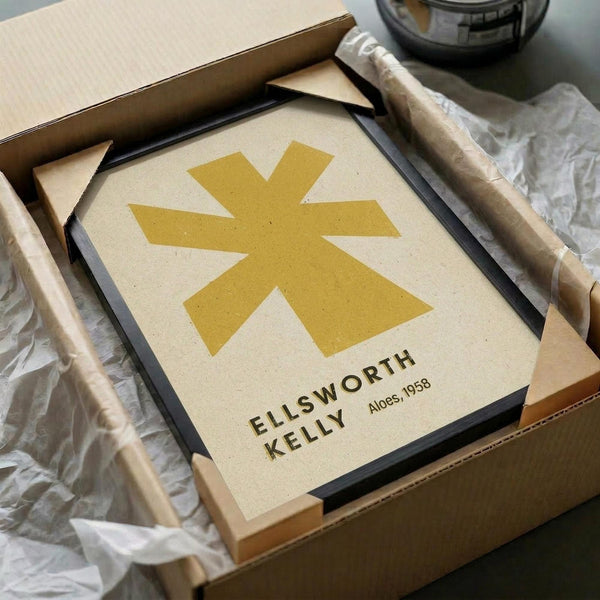May include: Framed abstract art print featuring a golden yellow geometric star or asterisk shape on cream background, displayed in black frame inside cardboard shipping box with protective tissue paper wrapping