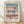 May include: Framed Bauhaus-style architectural poster print leaning against white textured wall on light wood floor. Features minimalist geometric building design in muted pink, sage green, and cream tones with clean lines and modern typography. Natural wood frame with cream matting, soft shadows cast across wall from natural lighting.