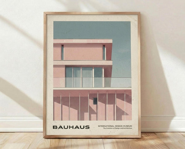 May include: Framed Bauhaus-style architectural poster print leaning against white textured wall on light wood floor. Features minimalist geometric building design in muted pink, sage green, and cream tones with clean lines and modern typography. Natural wood frame with cream matting, soft shadows cast across wall from natural lighting.
