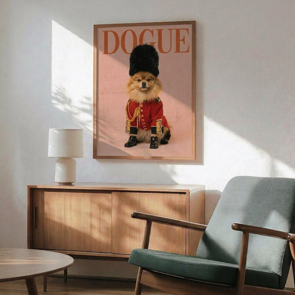 May include: Framed poster on pink background showing a Pomeranian dog wearing a red British Royal Guard uniform with gold details, black tall hat, and small boots. The poster displays "DOGUE" text at top in red letters. Scene shows modern living room with sage green mid-century armchair, wood sideboard, white ceramic lamp, and natural sunlight casting shadows on white walls.