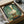 May include: Whimsical artwork of a white duck wearing a toilet paper roll on its head like a hat, painted in soft brushstroke style with green textured background. The framed piece is being carefully unpacked from a cardboard shipping box with protective tissue paper wrapping.