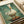 A whimsical painted illustration of a white duck standing on a wooden log, wearing a roll of toilet paper balanced on its head like a hat. The duck has an orange-yellow bill and orange webbed feet, set against a soft teal-green watercolor background. The artwork appears to be printed on canvas or textured paper with a cream border, photographed at an angle with white ribbon visible in the corner.