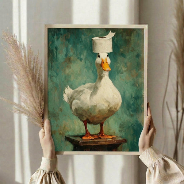 May include: Hands holding a framed art print of a white duck balancing a toilet paper roll on its head, standing on a wooden platform against a textured teal-green background. The whimsical painting shows the duck with orange beak and feet in a realistic style. Person wears cream-colored clothing with ruffled sleeves, and dried pampas grass is visible to the left against soft neutral curtains.