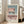 May include: Framed architectural poster with minimalist pink and teal building facade featuring geometric windows and balconies, displayed on wooden floor against white wall next to beige sofa in bright living room with natural window light
