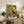 May include: Framed vintage-style poster featuring a black heron silhouette against an abstract painted background in warm yellows, oranges, and greens. The frame sits on a rustic wooden console table alongside books, pottery, and a fern plant, with a beige upholstered chair visible in a cozy living room setting with neutral walls and soft natural lighting.
