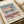May include: Minimalist architectural poster print displayed on cream colored paper with soft natural lighting. The artwork features a modern building rendered in muted pink, coral, and sage green tones with clean geometric lines and vertical slat details. Text reads "BAUHAUS" and "INTERNATIONAL DESIGN MUSEUM" in black typography. Print appears to have a vintage aesthetic with subtle texture. Shown alongside neutral fabric and warm ambient setting.