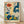 May include: Framed art print leaning against white wall on light wood floor. Poster features stylized botanical illustration with vibrant flowers and plants in retro folk art style. Design shows orange daisy, blue polka-dotted leaves, yellow wildflowers, green foliage, and purple blooms on cream background. Text reads "OSAKA '82" and "FLOWER MARKET" at bottom. Natural wood frame with thin border. Soft shadows cast on textured white wall.