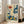 May include: Framed vintage-style poster featuring colorful botanical illustration with stylized flowers, leaves, and plants in navy blue, orange, yellow, and green on cream background. Text reads "Osaka '82" and "Flower Market" in Japanese and English. Light wood frame leaning against white wall next to beige sofa in bright, minimalist living room with large windows.